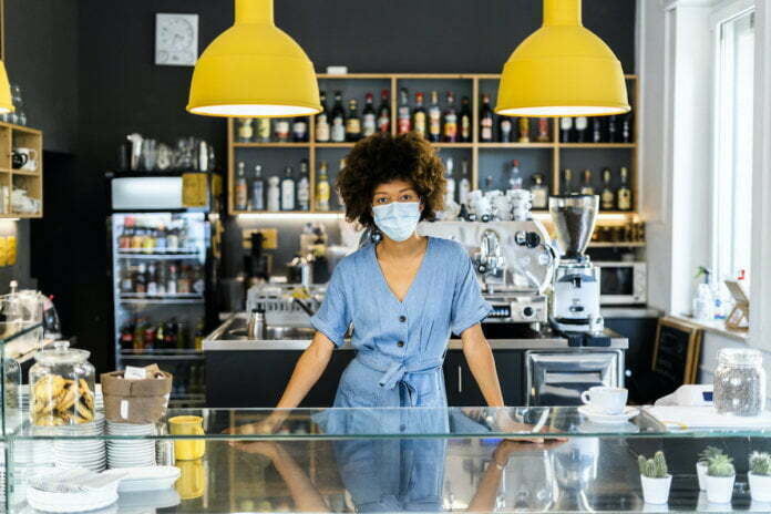 Young female owner wearing mask standing at counter in coffee shop
